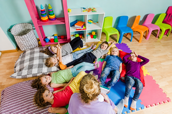 A teacher sits on a colorful foam mat reading to a group of seven diverse children lying on the floor in a bright classroom.