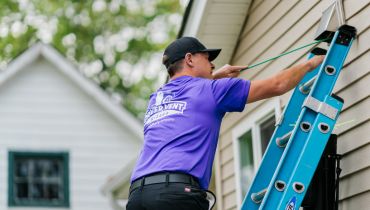 A worker in a purple shirt and black cap standing on a blue ladder.